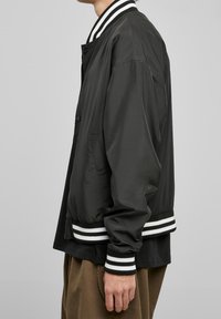 Czarna kurtka bomberska z lekkiego materiału, z paskowanym kołnierzem i mankietami, o luźnym kroju i bocznych kieszeniach.