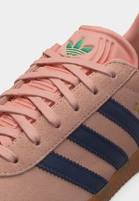 Ροζ σουέντ αθλητικό παπούτσι με ναυτικό μπλε ρίγες, ροζ κορδόνια και πράσινο λογότυπο Adidas στη γλώσσα, με χείλος σόλας από καουτσούκ.