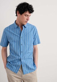 Camisa de botones de manga corta en azul claro con rayas verticales blancas, con un bolsillo en el pecho y botones grises. Tejido suave con un ajuste relajado.