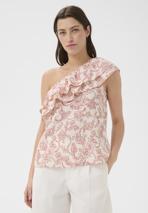 Femme portant un chemisier sans manches à une épaule avec imprimé paisley rose et détail volanté, associé à un pantalon blanc, debout devant un fond uni.