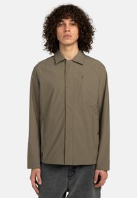 Khaki jacket with a pointed collar, front zipper, and chest pocket. Features smooth fabric and contrasting stitching.