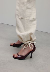 Pantalons beiges à cordon de serrage en tissu léger avec des chevilles froncées, associés à des sandales à talons hauts bordeaux brillantes et à bout ouvert.
