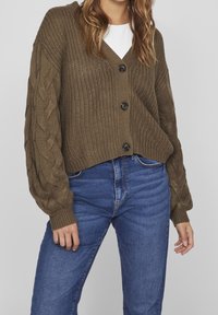 Kvinna som bär en brun grovstickad kofta med kabelärmar över en vit topp och blå högmidjade jeans mot en enfärgad bakgrund.