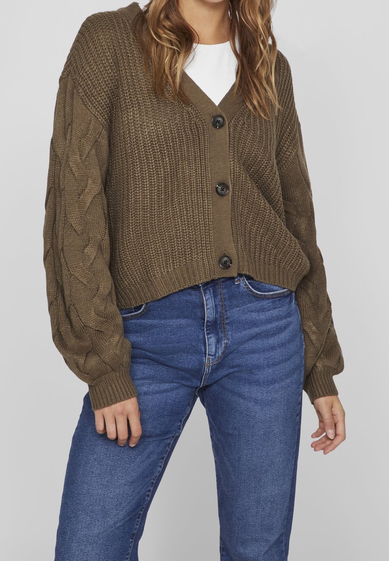 Kvinna som bär en brun grovstickad kofta med kabelärmar över en vit topp och blå högmidjade jeans mot en enfärgad bakgrund.