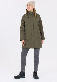 Olivgrön vattentät parkas med hög krage, dragsko i huven och två framfickor. Bärs över grå åtsittande jeans och svarta kängor.