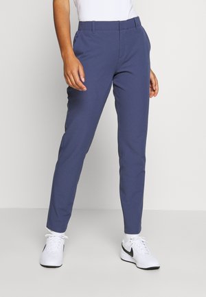 Personne portant un pantalon bleu marine slim et des baskets blanches Nike, se tenant devant un fond clair uni.