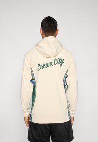 Krémová mikina s kapucňou so zeleným nápisom "Cream City" na chrbte. Má bočné zelené a modré akcenty. Vyrobená z hladkého, ľahkého materiálu.