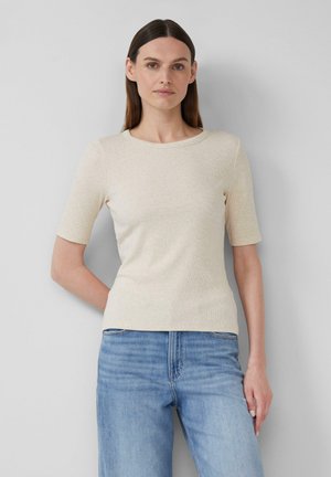 Een vrouw met lang bruin haar staat tegen een grijs muur, in een lichtbeige kortoog t-shirt en blauwe jeans, met haar handen op haar heupen.