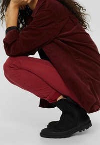 Bottines noires avec semelle en caoutchouc texturée, associées à une veste en velours côtelé bordeaux et à un jean skinny bordeaux, présentées en position accroupie.