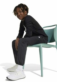 Zwart langemouwen shirt met een wit logo, zwarte broek en witte sneakers. Het onderwerp zit op een lichtblauwe stoel.