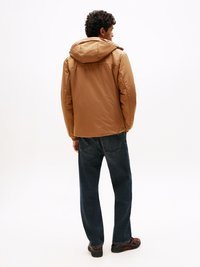 Giacca piumino marrone con cappuccio, caratterizzata da una texture liscia e opaca. Abbinata a jeans blu scuro e calzature nere. Vista posteriore.