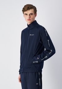 Navy blue track jacket in lightweight fabric, features a high collar, side pockets, and white branded accents along sleeves.