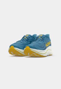 Sapatilhas desportivas azuis com parte superior em malha, detalhes amarelos e solas brancas espessas com um fundo em borracha texturizada. Nome da marca "HOKA" visível.