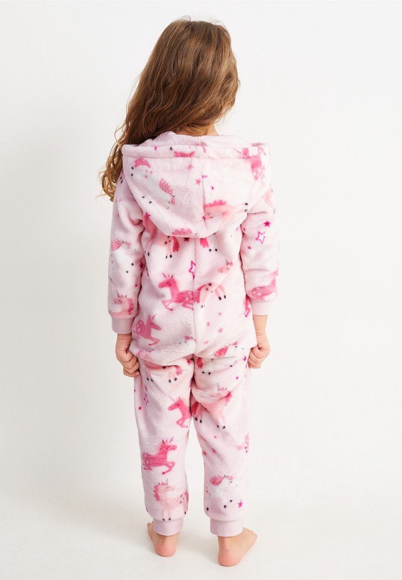Enfant aux longs cheveux portant un pyjama à capuche imprimé licornes roses, debout pieds nus de dos sur un fond uni.