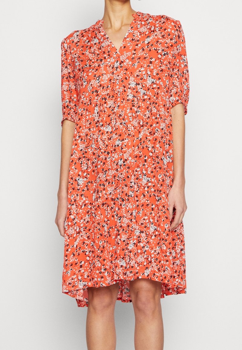Femme portant une robe rouge ample, longueur genou, avec un petit motif floral blanc et noir et des manches mi-longues.