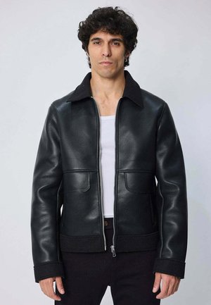 Homme aux cheveux bouclés foncés portant une veste en cuir noire par-dessus une chemise blanche, debout devant un fond uni.