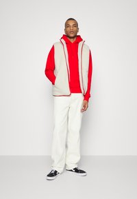 Gilet en polaire crème avec des bordures rouges sur un sweat à capuche rouge, associé à un pantalon blanc texturé et des baskets noires. Design à jambes droites plates.