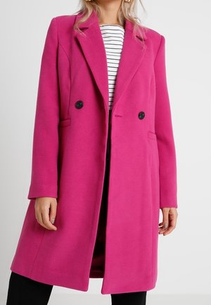 Kobieta w jasnoróżowym, dwurzędowym płaszczu, na czarno-białej bluzce w paski i czarnych spodniach, na jednolitym jasnym tle.