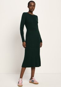 Frau steht in einem dunkelgrünen, gerippten Midikleid mit langen Ärmeln, kombiniert mit rosa und türkisfarbenen Sneakers, vor einem einfarbigen Hintergrund.