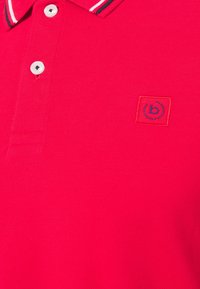 Polo rouge avec boutons blancs, col rayé blanc et marine, et petit logo "b" brodé en marine dans un carré sur la poitrine.