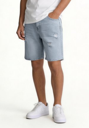 Man wearing light blue ripped denim shorts, white sneakers, and a white t-shirt, standing with one hand in pocket.