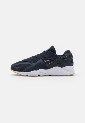 Mørkeblå Nike-sneaker med mesh- og ruskindsdetaljer, hvid mellemsål, lysebrun ydersål og sort plastik hælstøtte set fra siden.