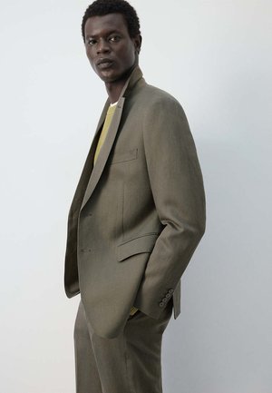 Homme portant un costume vert olive et un pull jaune clair, debout avec une main dans la poche sur un fond uni clair.