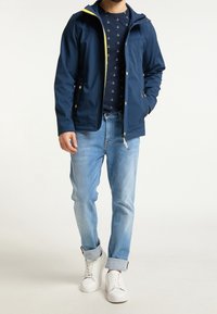 Navy jacka med gula detaljer, dragkedja och huva, bärs över en marinblå skjorta med ankarmönster. Ljusblå jeans och vita sneakers fulländar looken.