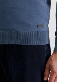 Blauer Pullover mit geripptem Saum und Bündchen, aus einem glatten Strickstoff gefertigt. Mit einem kleinen Metalldetail-Logo-Patch in der Nähe des Saums. Dunkle Hose sichtbar.