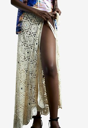 Inimene seob beeži hõlma külje, paljastades ühe jala, kandes värvikat ülaosa ja musti pahkluu rihmaga sandaale.