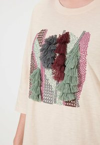 Cremfarbener T-Shirt mit strukturiertem Applikation, das grüne, bordeauxfarbene und graue Quasten zeigt, kombiniert mit verschiedenen gemusterten Stickdesigns.