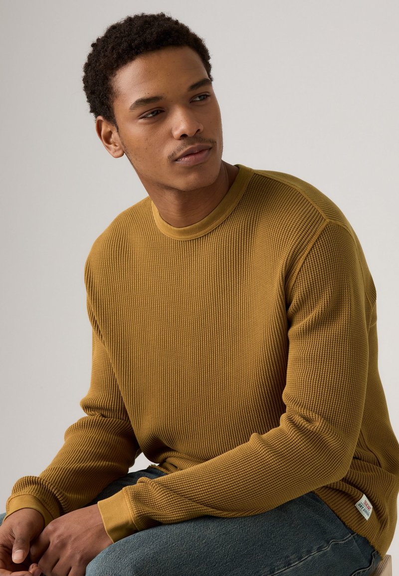 Senfgelbes geripptes Sweater aus strukturiertem Stoff, mit rundem Ausschnitt und engen Bündchen, kombiniert mit dunkelblauen Jeans.