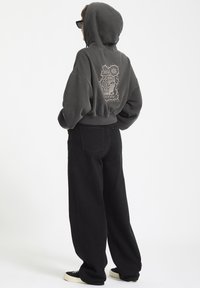 Donkergrijze cropped hoodie met een grafisch ontwerp op de achterkant, gecombineerd met losse zwarte broek en zwarte sneakers. Glad stoftextuur.