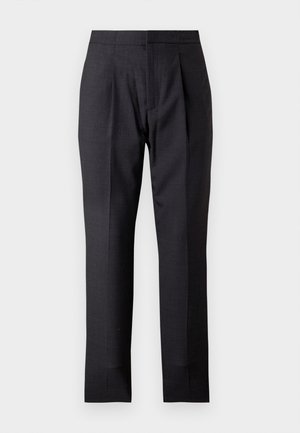 Homme portant un blazer beige sur une chemise bleu clair, un pantalon noir et des chaussures habillées noires, debout devant un fond blanc uni.