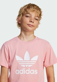 Pink cotton t-shirt featuring a large white Adidas logo with three stripes and leaf design on the front. Short sleeves and crew neck.
