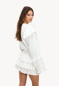 Witte jurk met een gestructureerde stof, ruches aan de mouwen en een gelaagde zoom. Kenmerken zijn rafelige randen en een getailleerde taille.