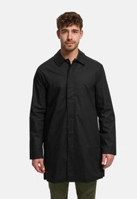 Pitkä musta takki, jossa on terävä kaulus, napitus ja suora leikkaus. Valmistettu sileästä materiaalista, siinä on pitkät hihat ja sivuhalkiot.