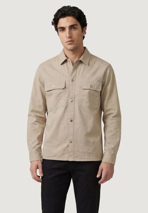 Jonge man die een beige overhemd met knopen draagt, voorzien van twee borstzakken, en zwarte broek, staand tegen een eenvoudige lichte achtergrond.