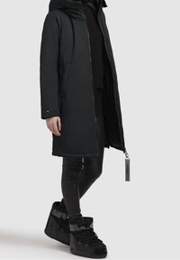 Parka negra acolchada con capucha, cierre de cremallera, bolsillos laterales y una etiqueta de acento. Combinada con leggings negros de corte slim y zapatillas altas a juego.