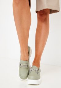 Ljust gröna loafers i texturerat konstläder, vita sulor och dekorativ metallhardware. Rund tå och slip-on-design.