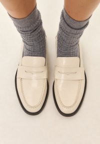 Weiße Lederloafers mit glänzender Oberfläche, ausgestattet mit einer schwarzen Gummisohle, getragen mit dicken, gerippten grauen Socken.