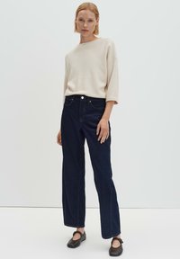 Donna che indossa un maglione crema a coste, jeans larghi scuri a gamba larga e scarpe nere Mary Jane, in piedi contro uno sfondo bianco semplice.