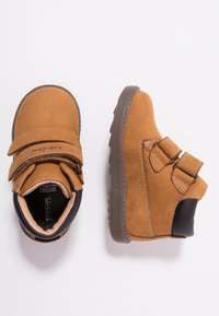 Braune Wildleder-Stiefeletten mit zwei Klettverschlüssen, einem schwarzen Lederakzent und einer strukturierten Gummisohle. Das Design mit Absatz bietet Stabilität.
