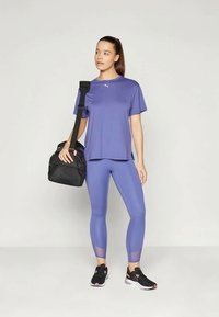 Top sportivo viola, leggings coordinati con accenti in rete, sneakers nere e borsa a tracolla nera. Tessuto liscio, vestibilità comoda.
