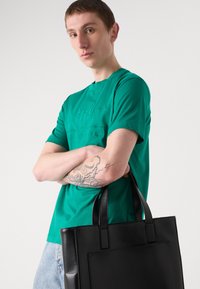 Juoda odinė rankinė su dviem rankenomis, lygiu paviršiumi ir stačiakampiu pavidalu. Modelis vilki žalią marškinėlį su apvalia iškirpte ir subtiliu prekės ženklu.