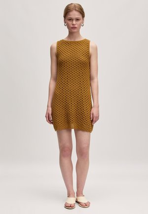 Strickkleid ohne Ärmel in senfgelb mit offenem, strukturiertem Muster. Gerader Schnitt und knielang, kombiniert mit hellen Sandalen.