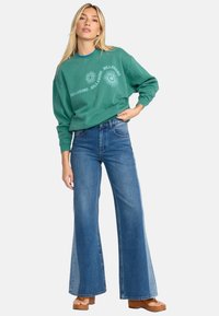 Groene katoenen sweatshirt met witte tekst en zonafbeeldingen, gecombineerd met blauwe flared jeans en bruine platform sandalen. Model staat met de hand op de heup.