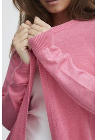 Roze gebreide cardigan met een geribbelde halslijn en manchetten, met een losse pasvorm. De stof lijkt zacht en textuurrijk te zijn.