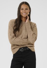 Lichtbruin gebreid sweater met lange mouwen, geribbelde manchetten en een ronde halslijn, gecombineerd met zwarte jeans en een zwarte riem.