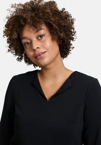 Schwarze Bluse mit V-förmigem Ausschnitt. Lange Ärmel, weicher Stoff, minimale Details und eine einzelne Brusttasche auf der linken Seite sichtbar.